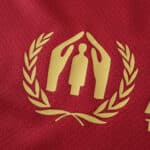 Red jersey with a gold emblem featuring two hands, a figure in the center, and laurel leaves on either side.