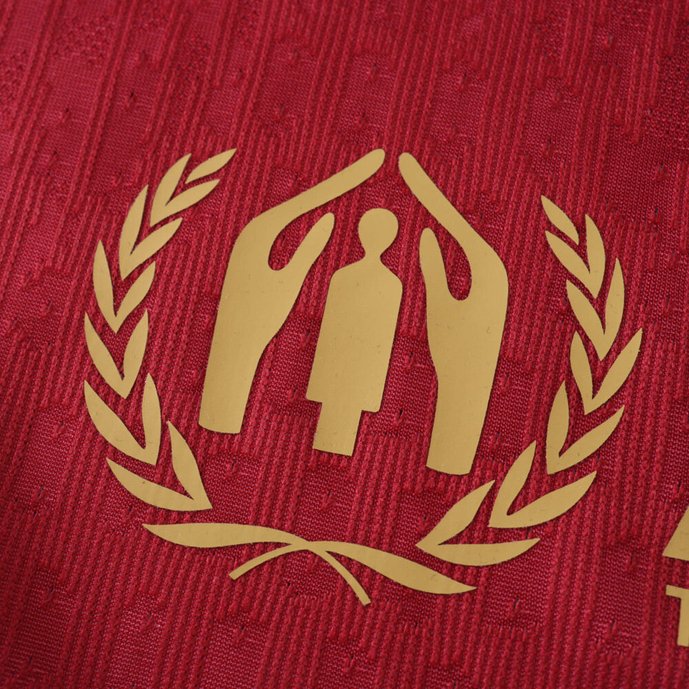 Close-up of a Barcelona x Coldplay special release player version jersey, featuring a gold emblem with laurel wreath and hands.
