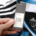 Close-up of a hand holding the tag on a Chelsea Retro Third 2011/12 Fan Version jersey showing size and care details.