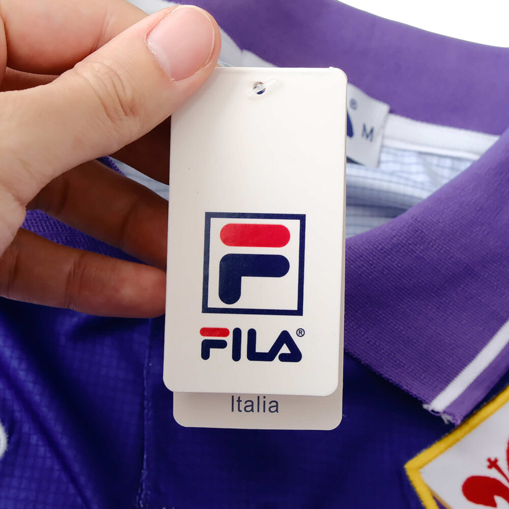 Close-up of a hand holding the FILA tag attached to the collar of Fiorentina Retro Home 1998/99 Fan Version jersey.