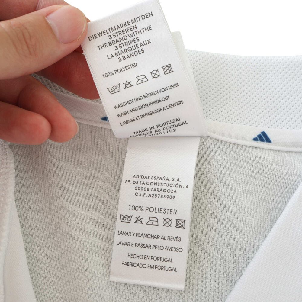 Close-up of the care and fabric tags on the Real Madrid Retro Home 2001/02 Fan Version jersey. Shows washing instructions and material details.