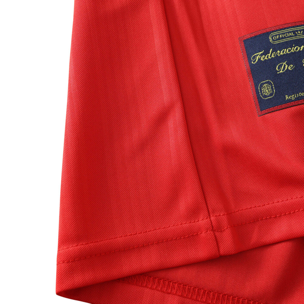 Close-up of the lower side of a red Spain Retro Home 1996 Fan Version jersey, showing the official badge.