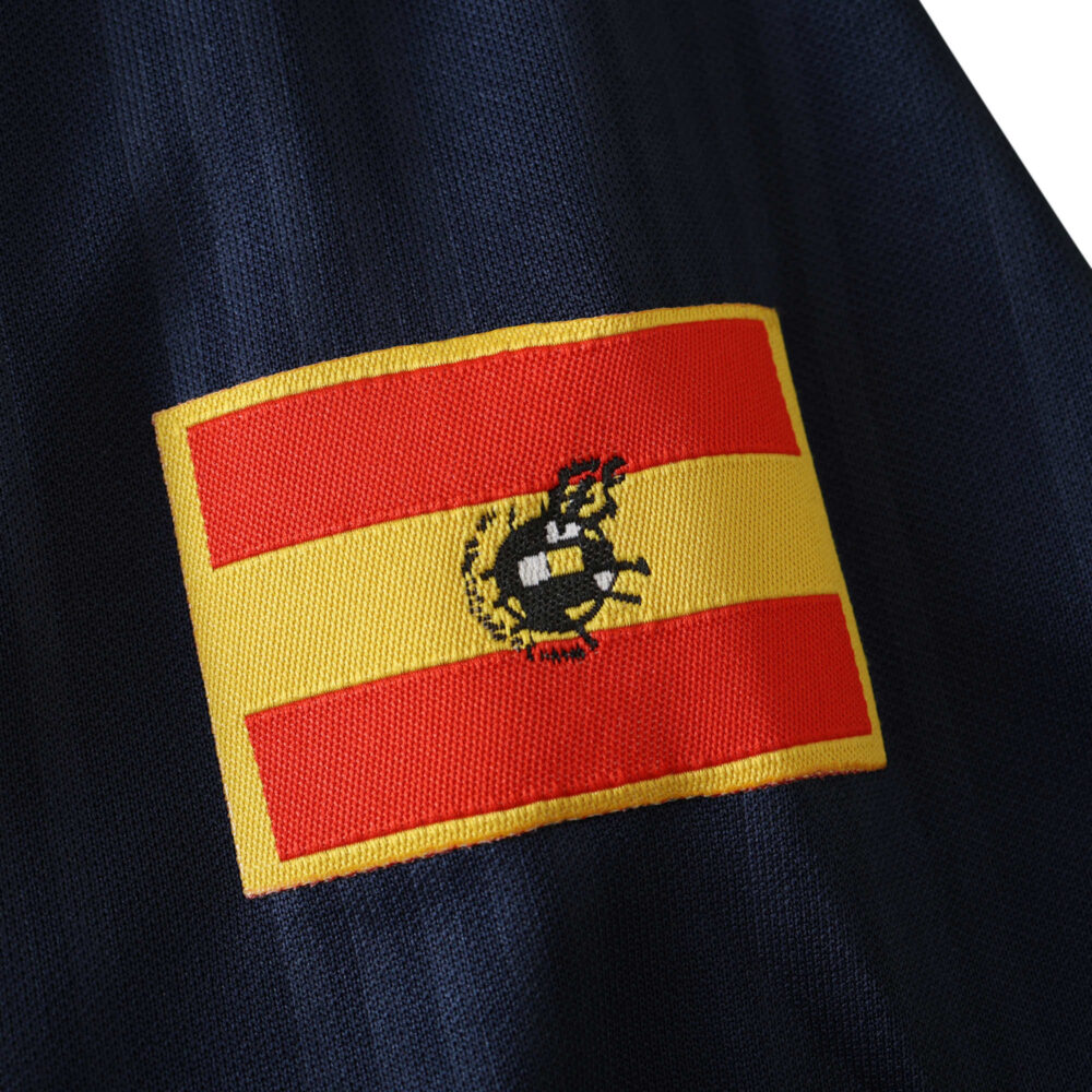 Close-up of the Spain Retro Home 1996 Fan Version soccer jersey, featuring an embroidered Spanish flag on navy fabric.