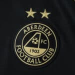 Close-up of Aberdeen FC 2025/26 away fan jersey badge with two gold stars on a dark background.