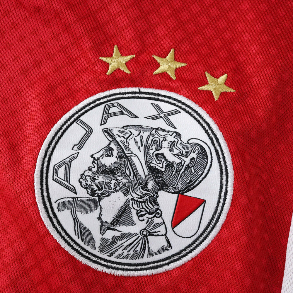Ajax Home 2025/26 Fan Version jersey badge with club crest and three gold stars on red fabric.