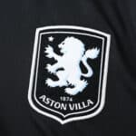 Close-up of the Aston Villa 2025/26 away fan jersey featuring the club's white lion crest on black fabric.