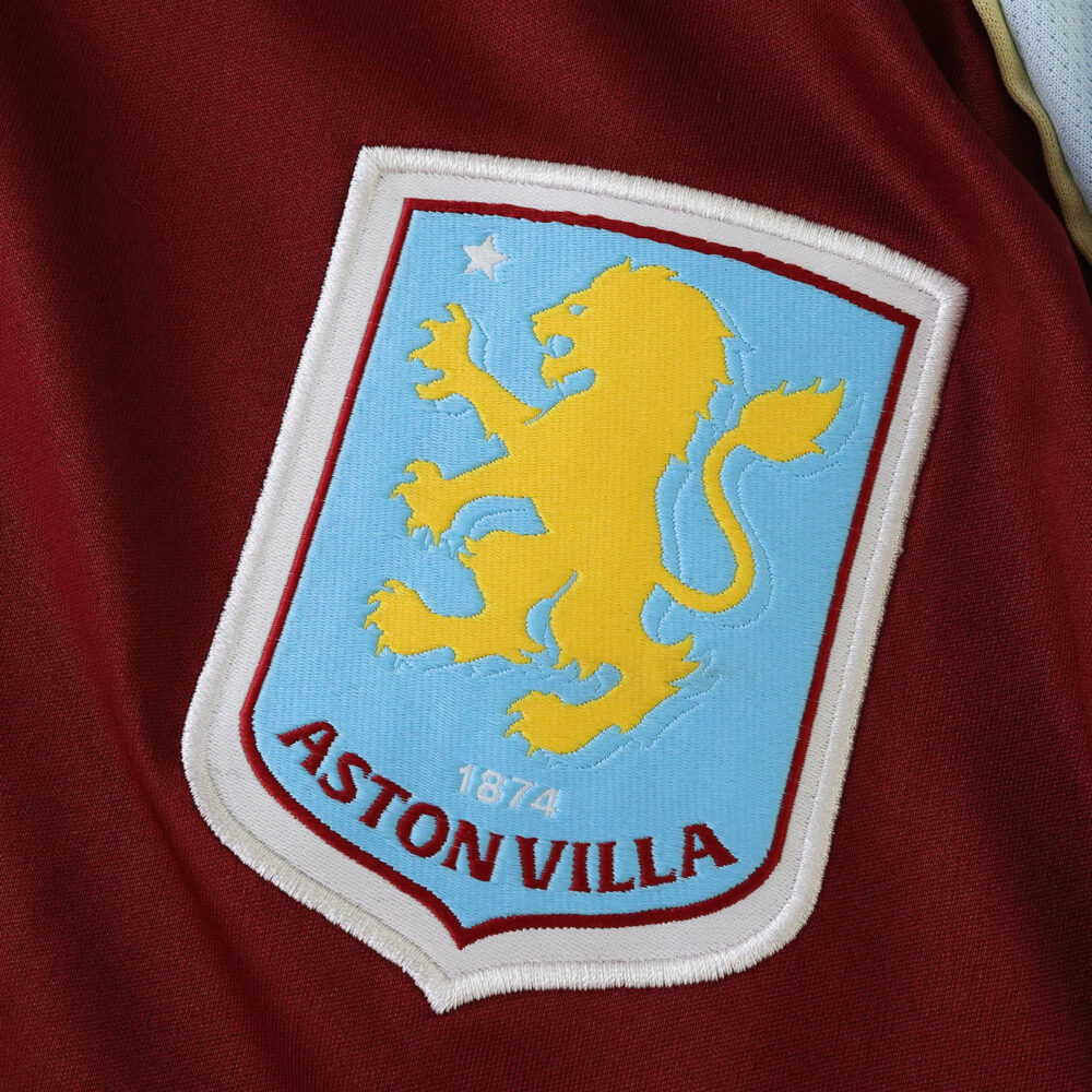Close-up of the Aston Villa crest on the 2025/26 home fan version jersey, featuring a yellow lion and blue shield.