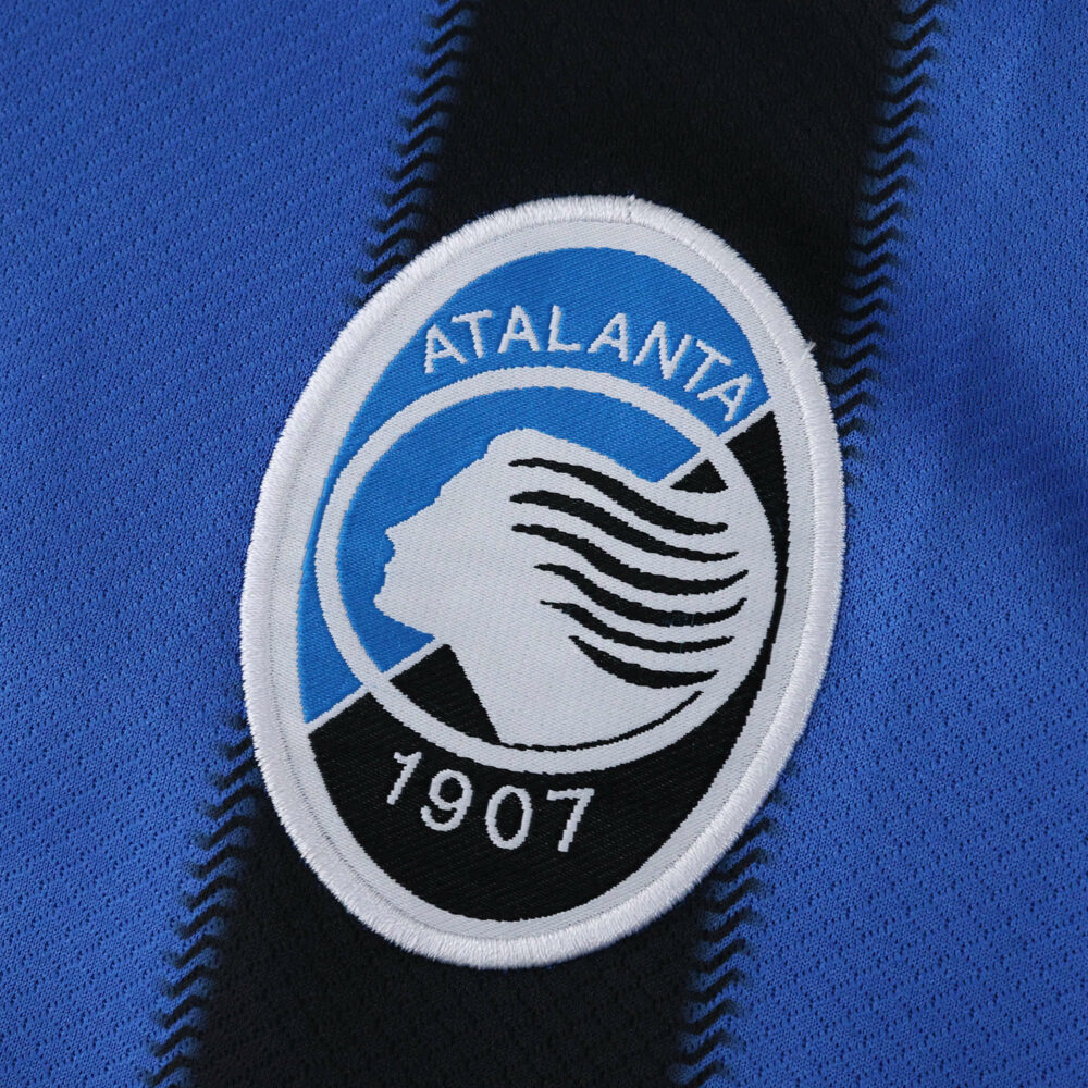 Close-up of the Atalanta 1907 crest on the 2025/26 home fan jersey, blue and black fabric visible.