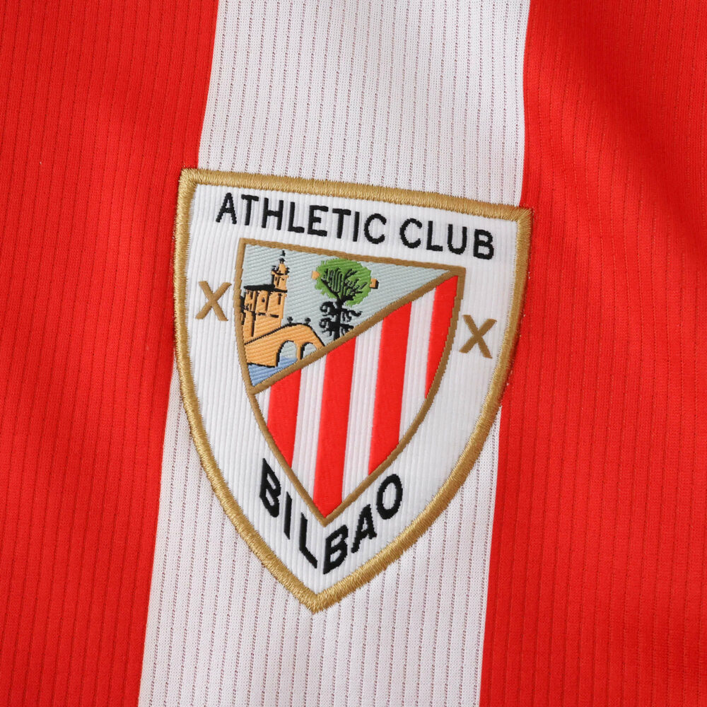 Close-up of the Athletic Bilbao 2025/26 home jersey badge on red and white striped fabric.