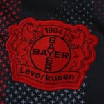 Close-up of the Bayer Leverkusen 2025/26 home fan version jersey crest on dark patterned fabric.
