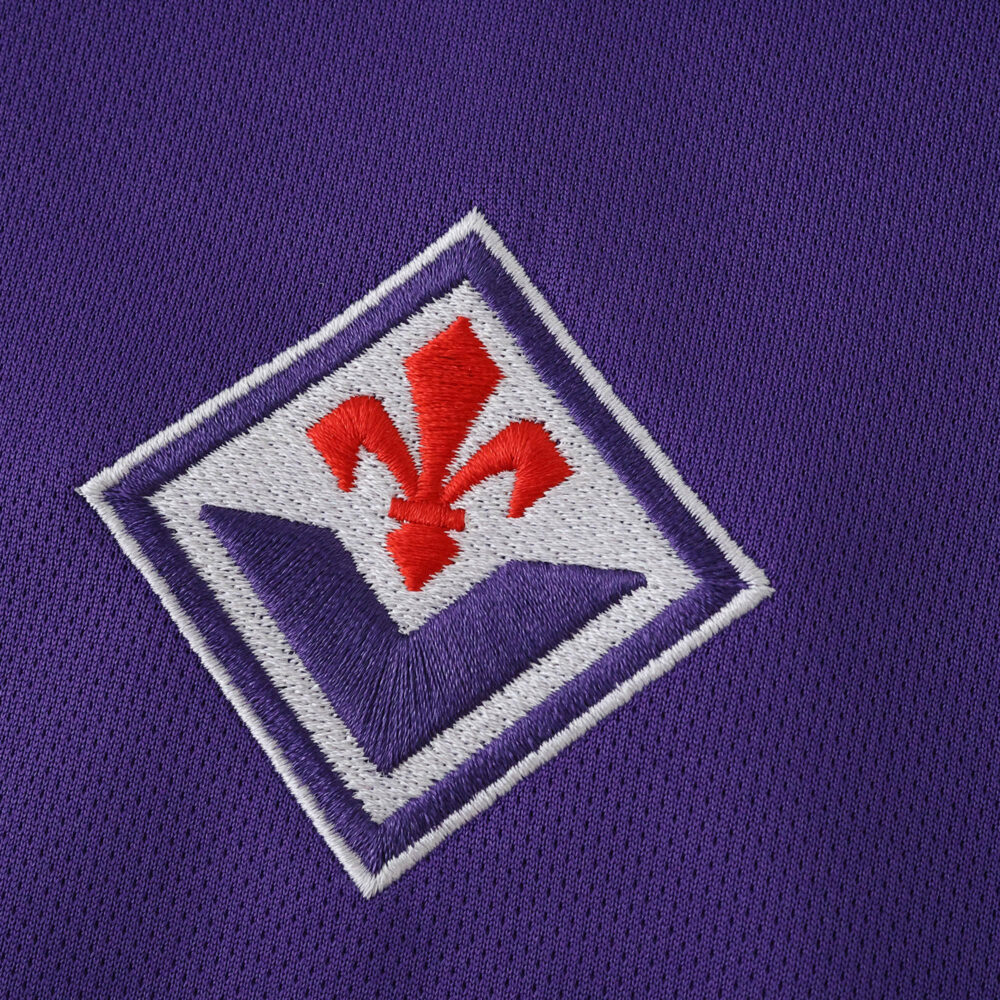 Close-up of the Fiorentina 2025/26 home fan jersey badge on purple fabric.