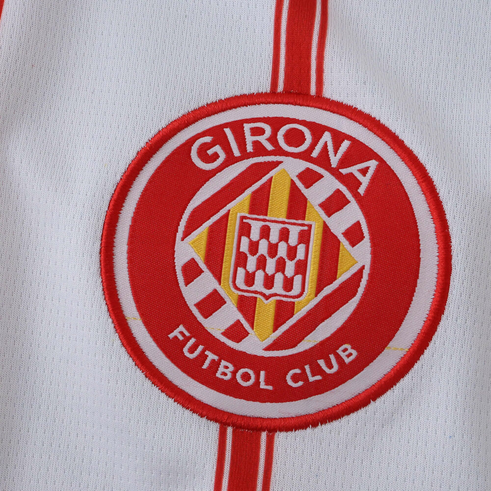Close-up of Girona FC 2025/26 home jersey badge with red and white club crest and vertical stripes.