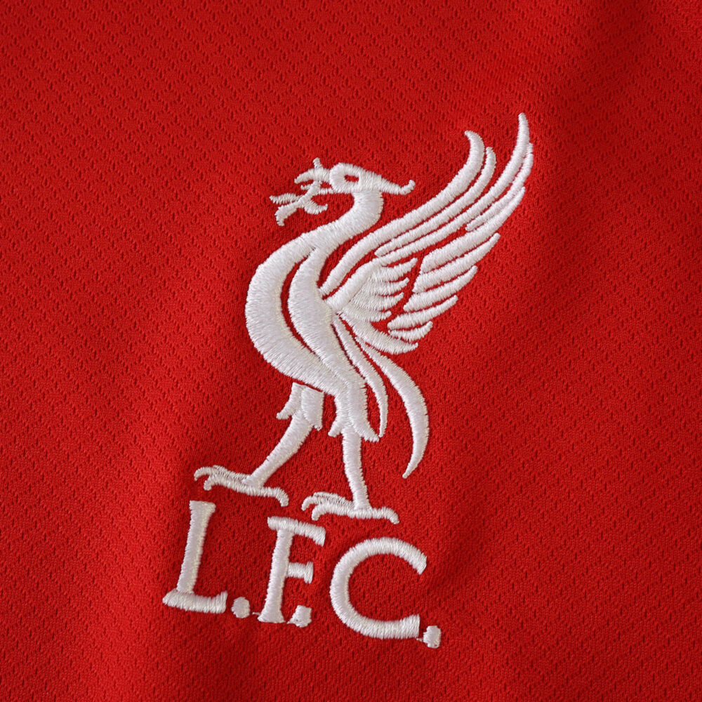 Close-up of the white Liverpool FC crest embroidered on the red 2025/26 home fan version jersey fabric.