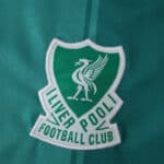 Close-up of the Liverpool Third 2025/26 Fan Version crest on a green football jersey.