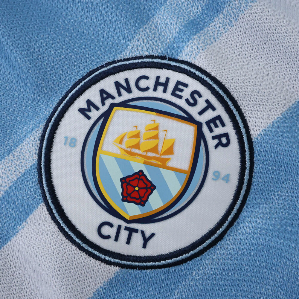 Manchester City 2025/26 Home Fan Version badge on light blue and white striped fabric.