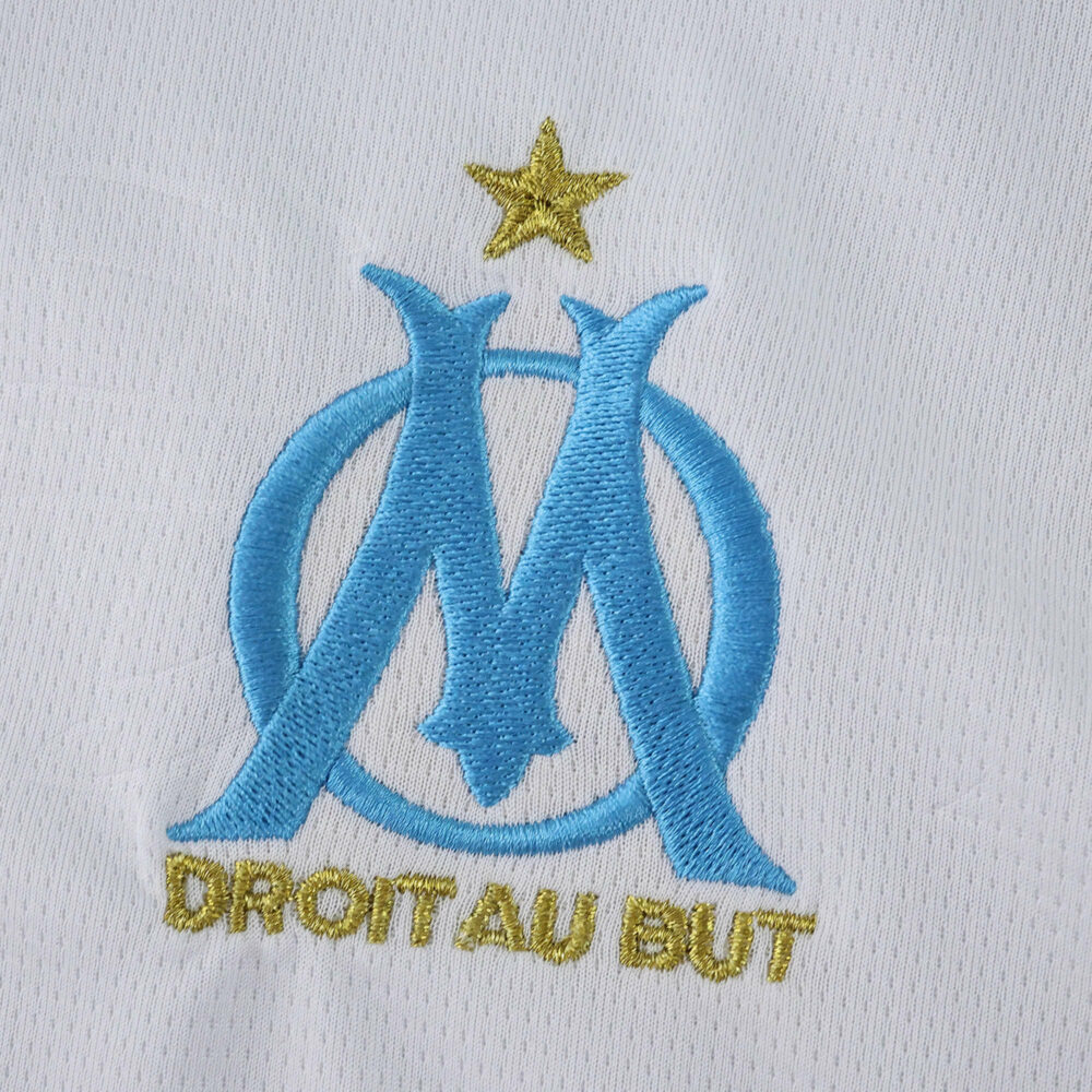 Olympique Marseille crest with gold star and "Droit Au But" on the 2025/26 home fan version jersey.
