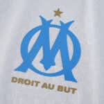 Olympique Marseille 2025/26 home jersey detail with blue crest and gold "Droit Au But" slogan.