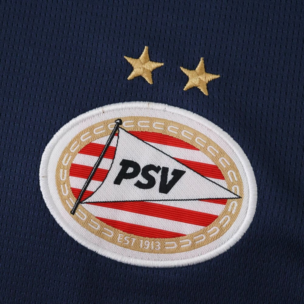 Close-up of the PSV Eindhoven 2025/26 away fan jersey badge with two gold stars above the crest.