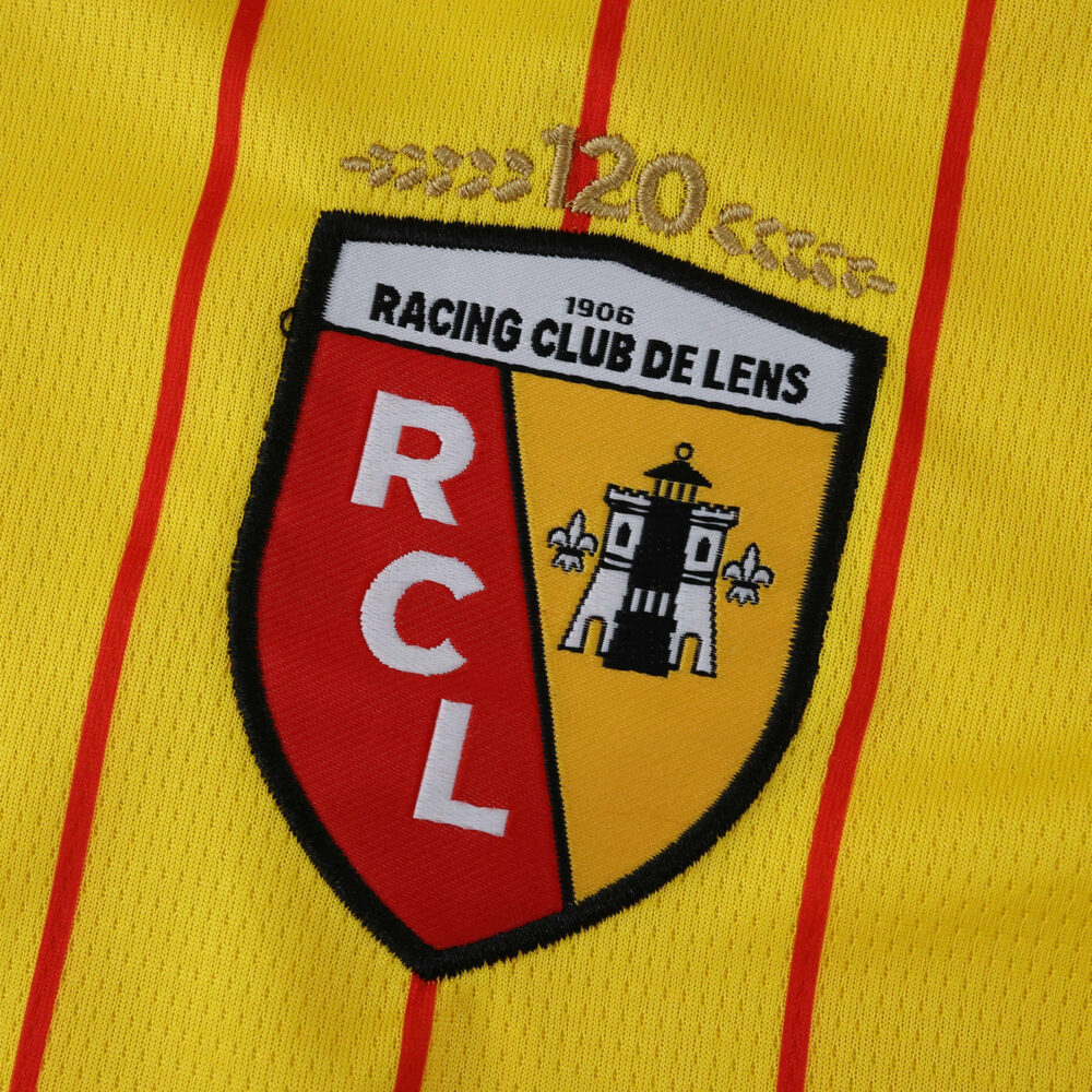 Close-up of RC Lens 2025/26 home fan jersey crest on yellow fabric with red stripes.