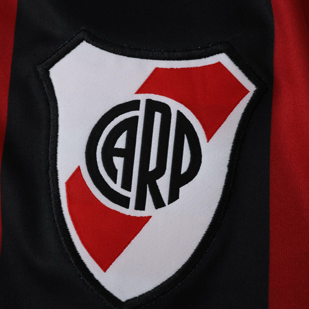 Close-up of the River Plate crest on the 2025/26 away fan version jersey with black and red stripes.