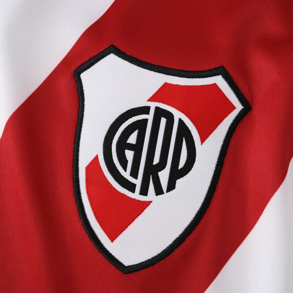 Close-up of the River Plate 2025/26 home jersey badge, featuring the club crest on red and white fabric.