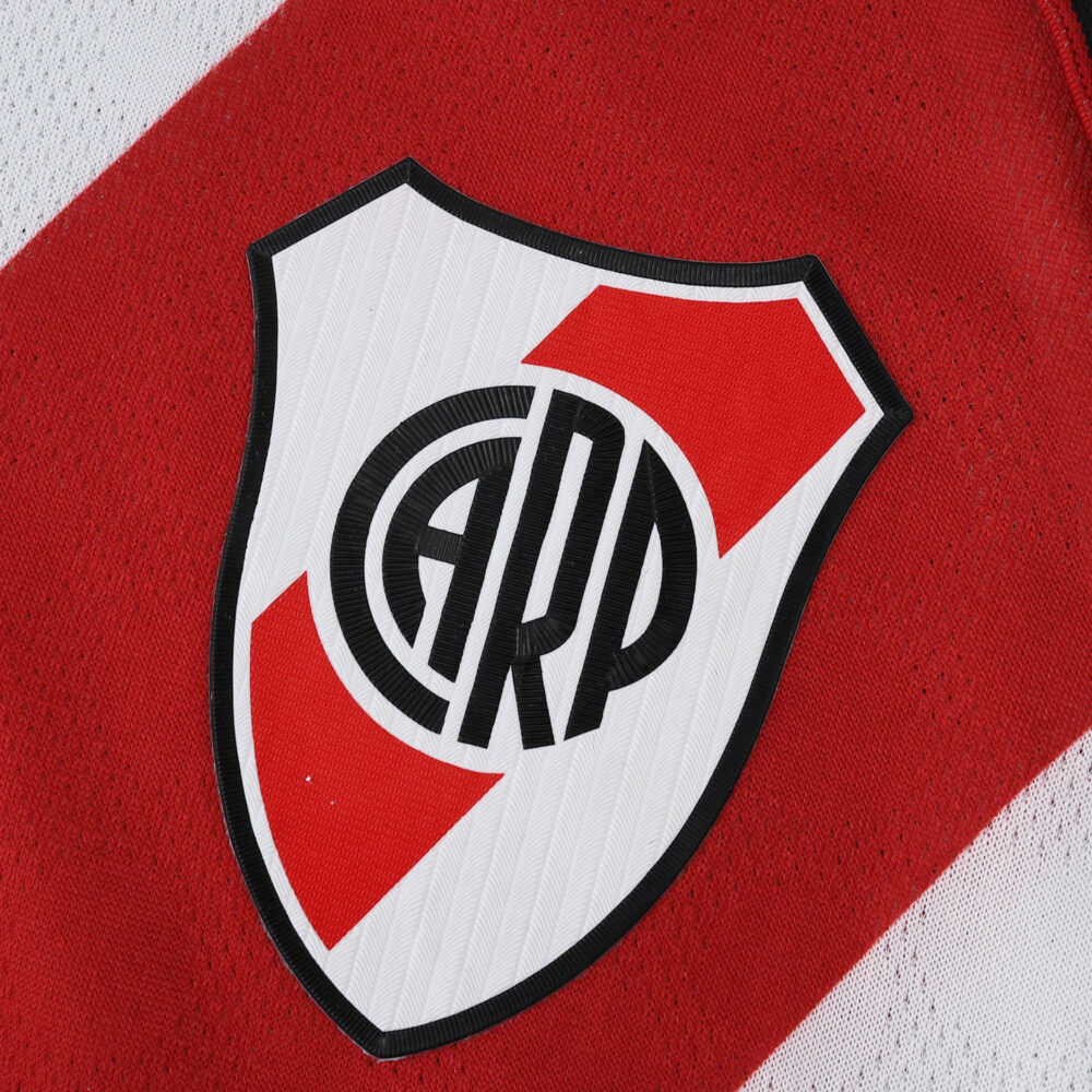 Close-up of the River Plate 2025/26 home jersey crest with red diagonal stripe and club logo.