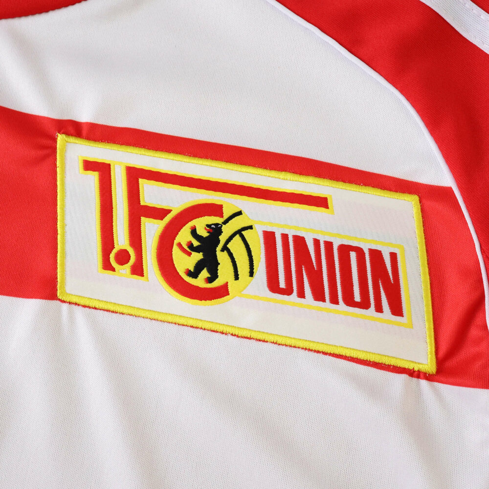 Close-up of the Union Berlin 2025/26 home jersey badge on white fabric with red details.