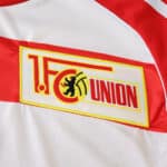 Close-up of the Union Berlin 2025/26 home jersey badge on white fabric with red details.