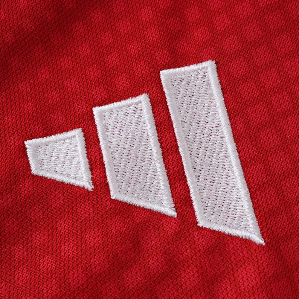 Close-up of the white Adidas logo stitched on the red Ajax Home 2025/26 Fan Version jersey fabric.