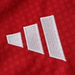 Close-up of the white Adidas logo stitched on the red Ajax Home 2025/26 Fan Version jersey fabric.