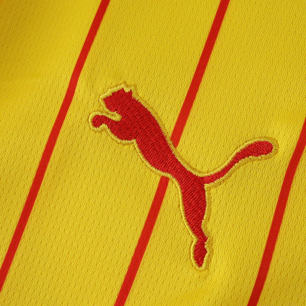 Close-up of the RC Lens 2025/26 home fan jersey featuring the red Puma logo on yellow fabric with red stripes.