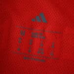 Close-up of Bayern Munich 2025/26 home player version jersey size tag and Adidas logo on red fabric.