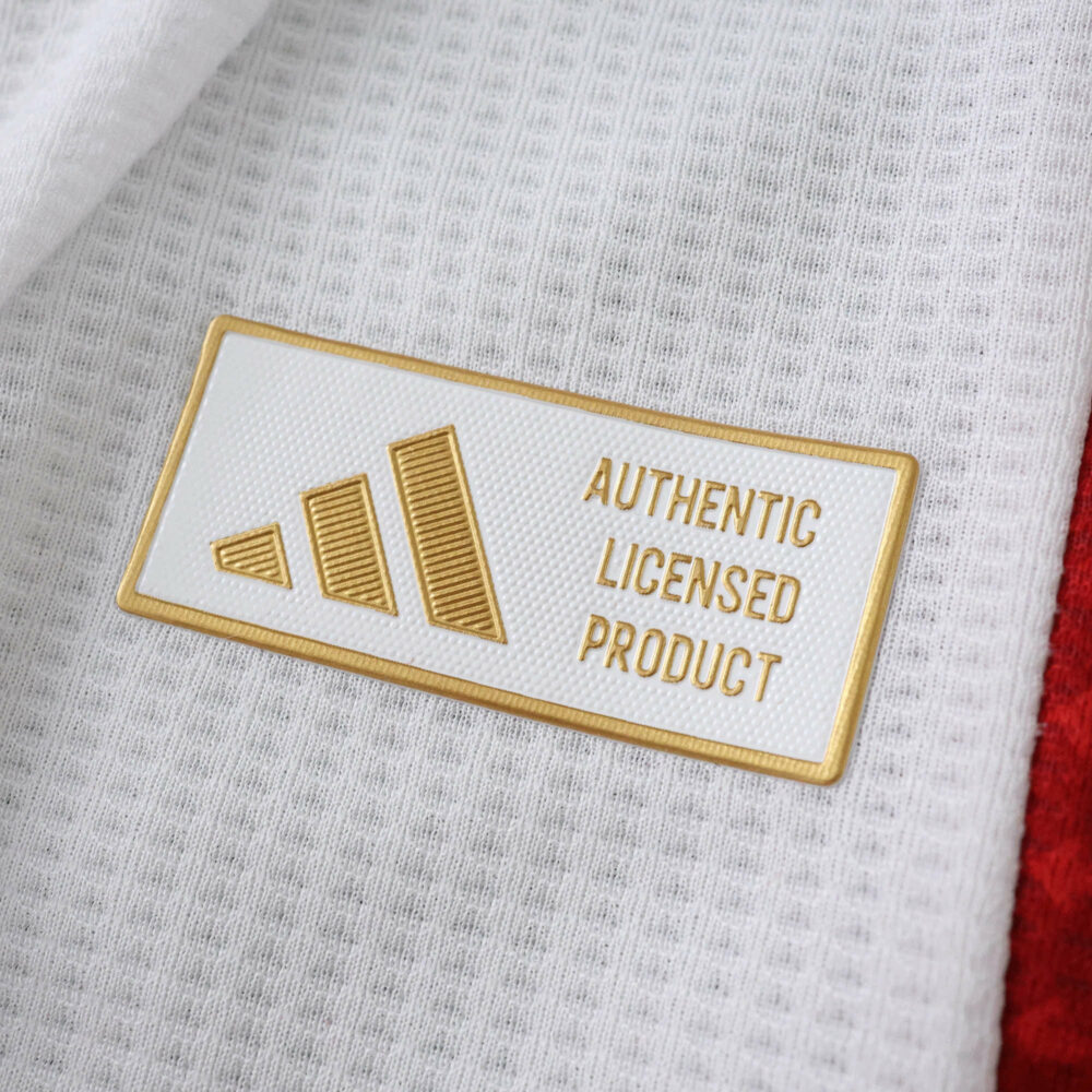 Close-up of the gold Adidas "Authentic Licensed Product" badge on the Ajax 2025/26 player jersey.