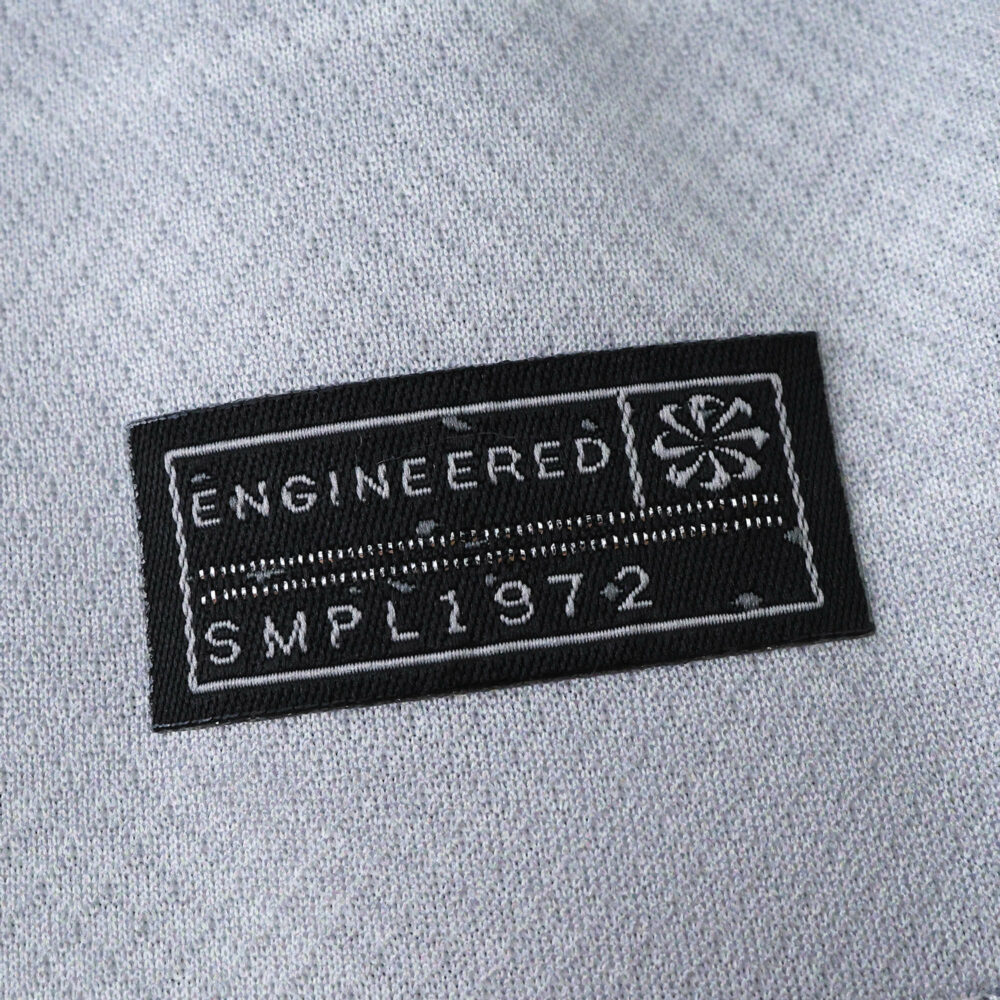 Close-up of the "Engineered SMPL 1972" tag on the VfL Wolfsburg Away 2025/26 Fan Version jersey.