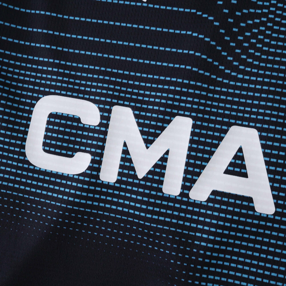 Close-up of "CMA" logo on Olympique Marseille 2025/26 Away Fan Version jersey with blue patterned background.