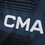 Close-up of "CMA" logo on Olympique Marseille 2025/26 Away Fan Version jersey with blue patterned background.