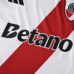 Close-up of River Plate 2025/26 home fan jersey featuring sponsor logo and red diagonal stripe.