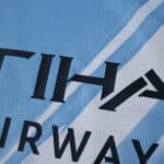 Close-up of the Manchester City 2025/26 home fan jersey showing sponsor text and light blue pattern.