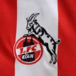 Close-up of the FC Köln crest on the 2025/26 home fan jersey, featuring a goat and red-white stripes.