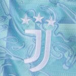 Close-up of Juventus Away 2025/26 Fan Version jersey featuring club crest with three stars on blue patterned fabric.