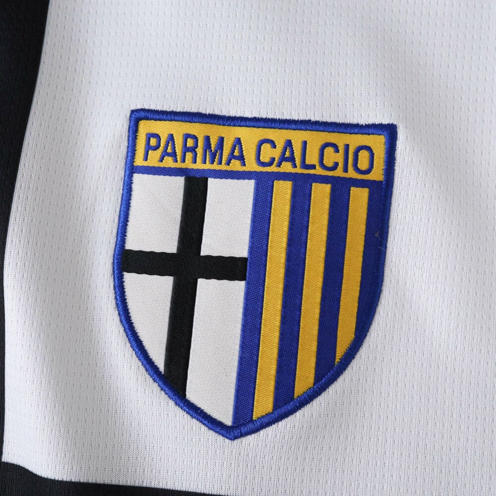 Parma Calcio badge on the 2025/26 home fan jersey, featuring black cross and yellow-blue stripes.