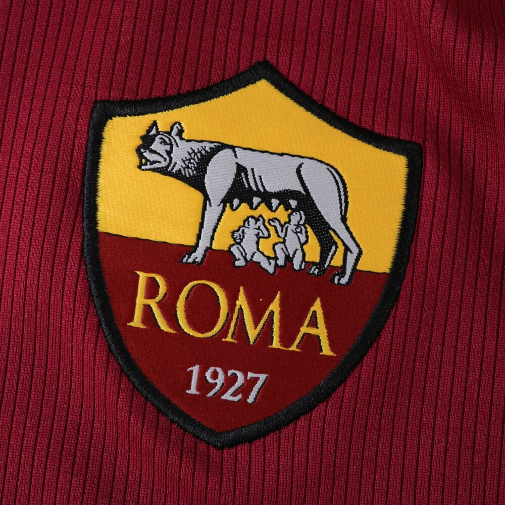 AS Roma 2025/26 home jersey badge featuring the she-wolf and twins emblem with "ROMA 1927" text.