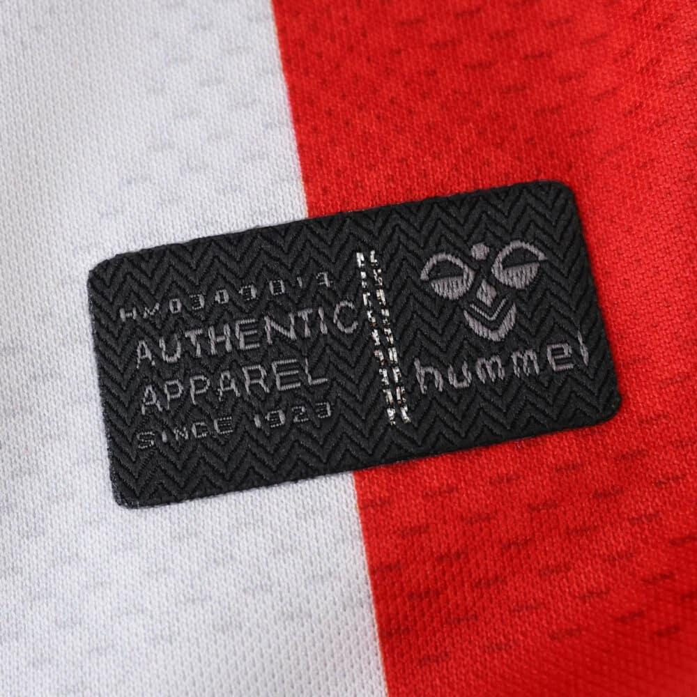 Close-up of the Hummel authenticity label on the FC Köln 2025/26 home fan jersey, red and white background.