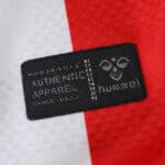 Close-up of the Hummel authenticity label on the FC Köln 2025/26 home fan jersey, red and white background.