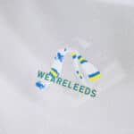 Close-up of Leeds United 2025/26 home fan jersey featuring "WE ARE LEEDS" text and blue-yellow scarf graphic.