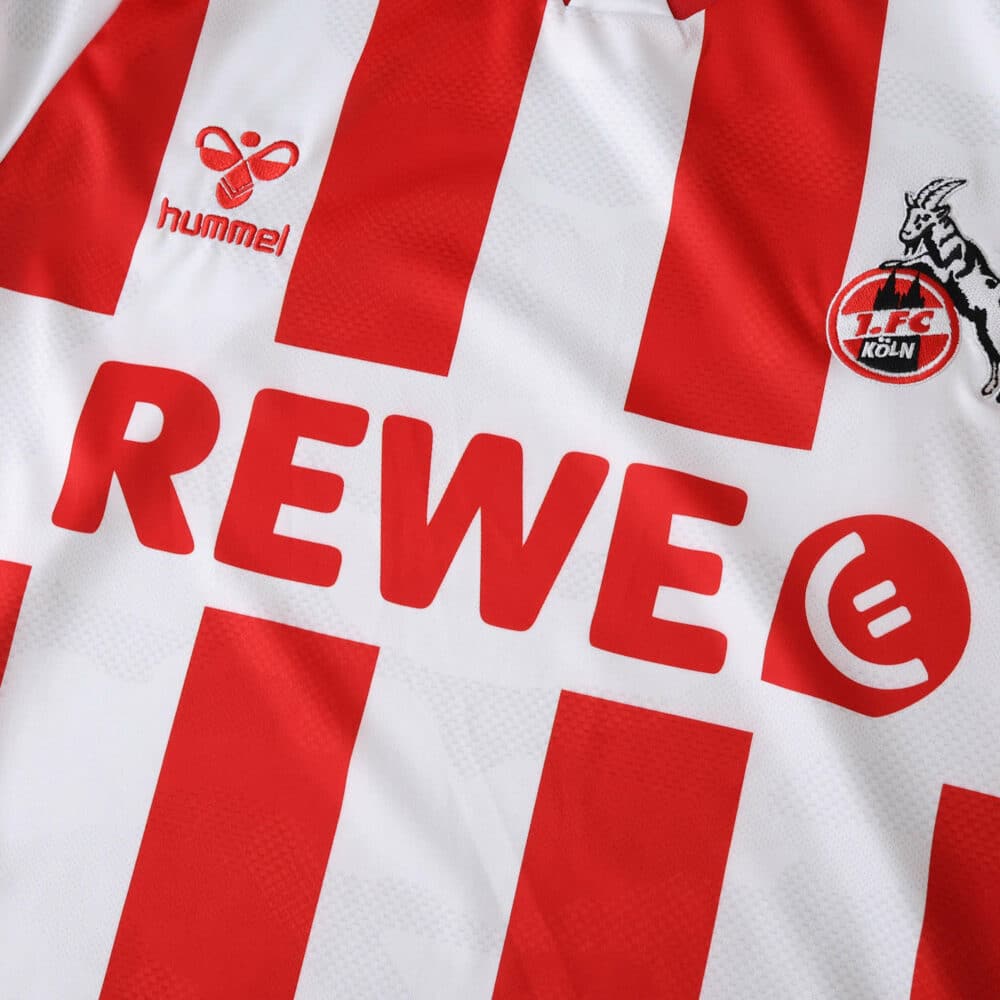 Close-up of FC Köln 2025/26 home fan jersey showing red stripes, club badge, Hummel and REWE logos.