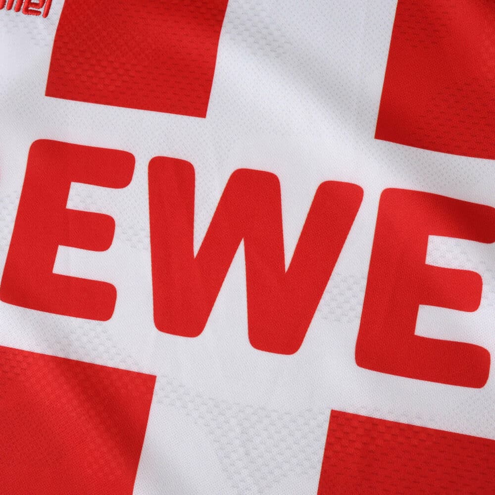 Close-up of the FC Köln 2025/26 home fan jersey showing the sponsor logo "REWE" on a red and white cross pattern.