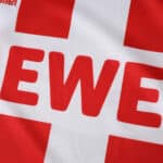 Close-up of the FC Köln 2025/26 home fan jersey showing the sponsor logo "REWE" on a red and white cross pattern.