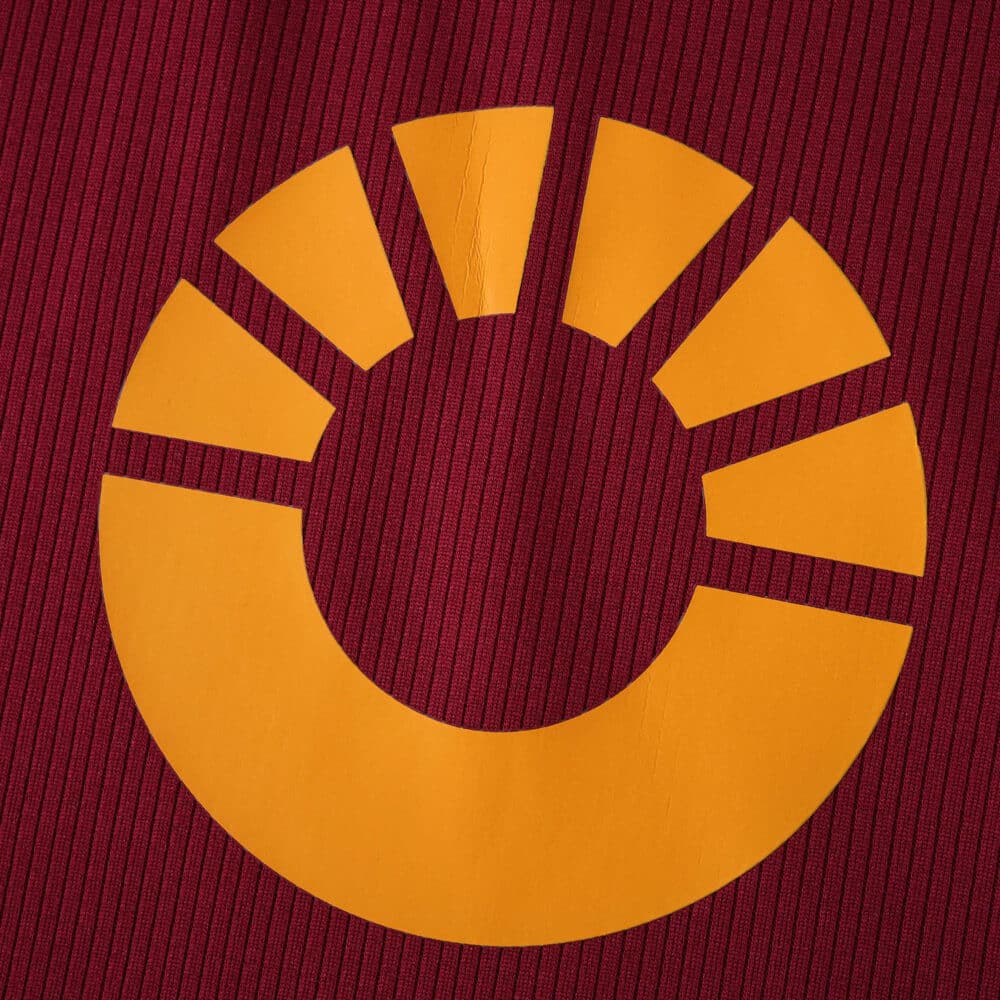Close-up of the yellow Roma 2025/26 home kit sponsor logo on a dark red fabric background.