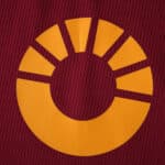 Close-up of the yellow Roma 2025/26 home kit sponsor logo on a dark red fabric background.